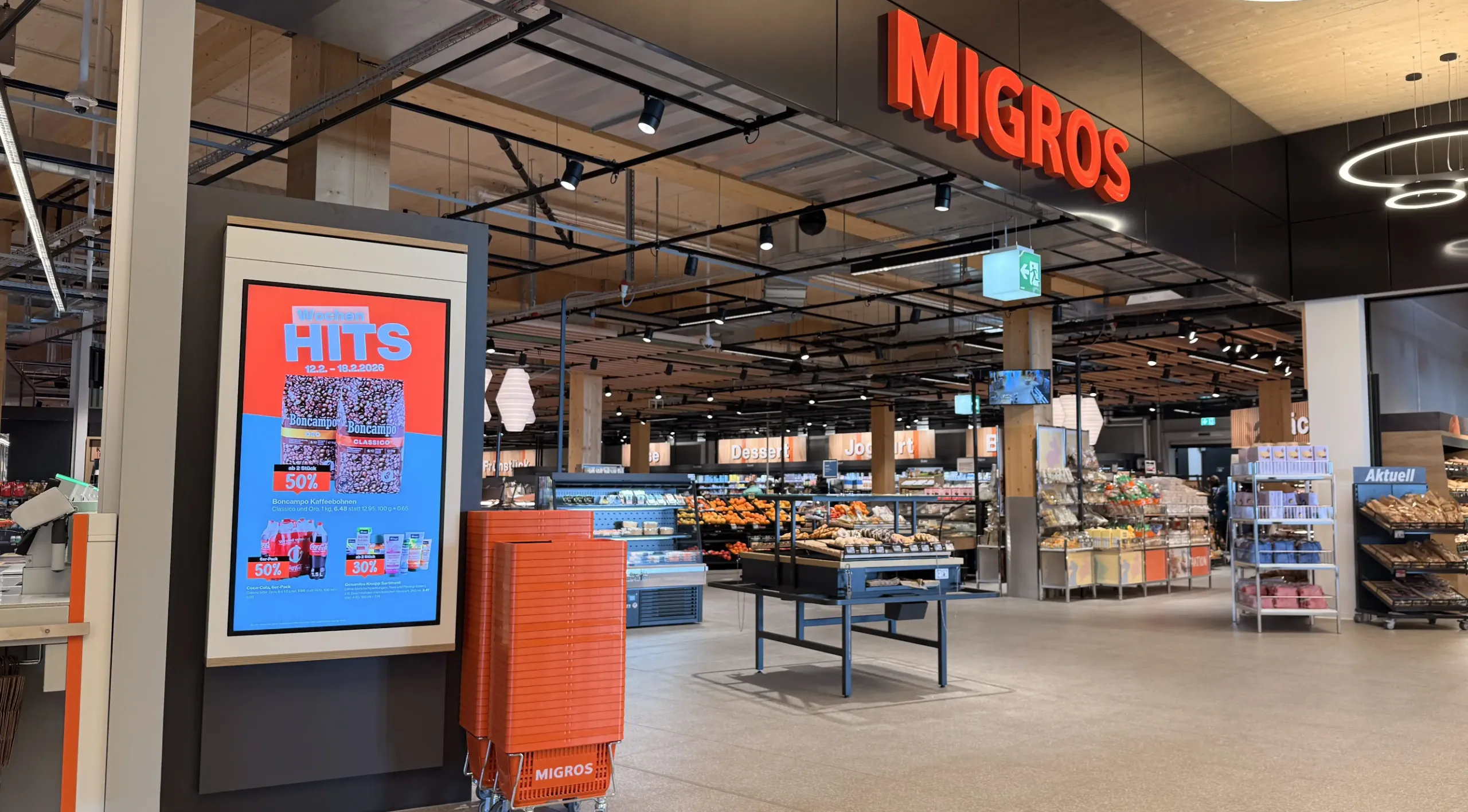 Eingang Migros Supermarkt mit grossem Digital Signage Display links und Migros Einkaufskörben daneben