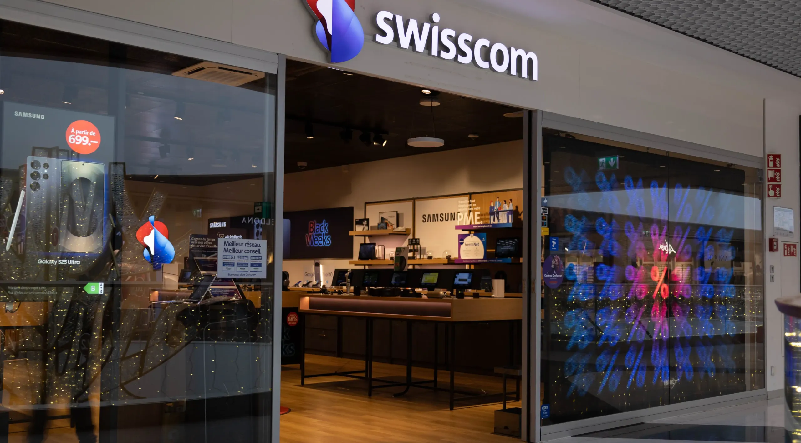 Shoppers passing a transparent shop window LED with Black Weeks advertising at the Swisscom store Geneva Balexert, implemented by jls.