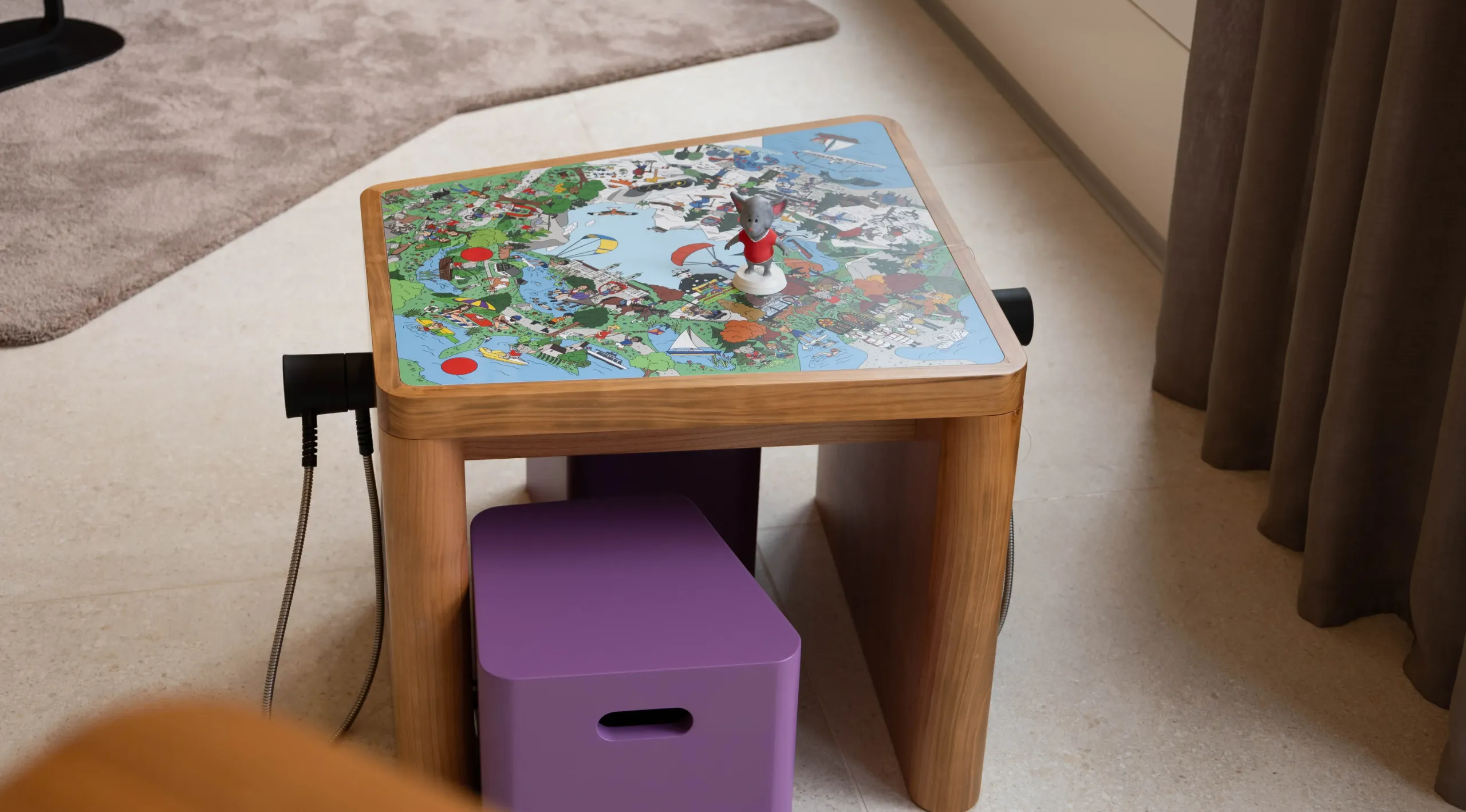 Nahaufnahme des illustrierten Wimmelbild-Tischs mit der Kabi-Figur und einem violetten Hocker darunter.
